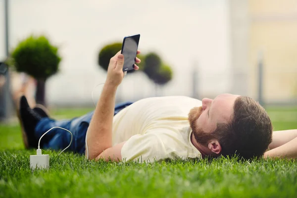 Natureza e tecnologia. Um homem com barba jaz em uma grama verde com um smartphone em suas mãos está cobrando banco de poder . — Fotografia de Stock