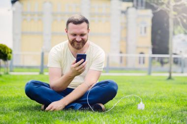 Doğa ve teknoloji. Bir sakallı bir adam yatıyor bir yeşil çim onun elinde bir akıllı telefon ile güç banka şarj.