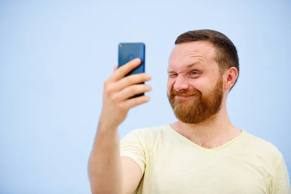 Veselý vousatý muž mrká na telefon, vhodný pro reklamu, na modrém pozadí — Stock fotografie