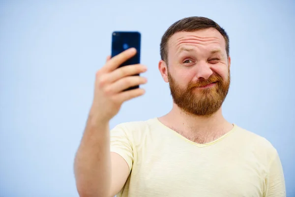 Veselý vousatý muž mrká na telefon, vhodný pro reklamu, na modrém pozadí — Stock fotografie