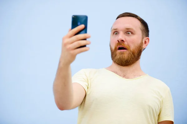 Muž s červenou bradku vypadá do telefonu s překvapením a extáze světlé letní foto na modrém pozadí — Stock fotografie