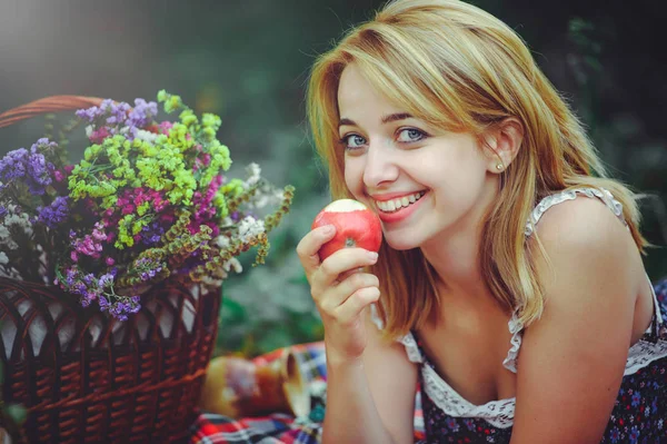 Vestidos de mujer feliz comer manzanas y mentiras sonrientes en la naturaleza, cerca de ramo de flores silvestres, estilo de vida saludable y relajación, cara linda y ojos azules — Foto de Stock
