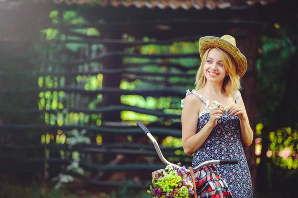 Mulher bonita com bicicleta na natureza, cesta com flores e floresta no fundo, foto alegre aconchegante em um chapéu — Fotografia de Stock