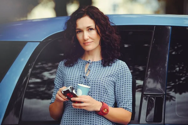 Hermosa mujer rizada bebiendo café cerca del coche en un dre azul — Foto de Stock