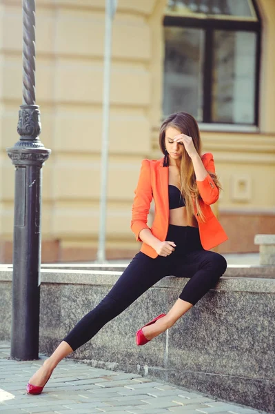 A sweet face is a mystery. Fashion portrait of a happy woman in red style jacket and black pants sits on the background of new houses, a new building. place for banner and advertising — Stock Photo, Image