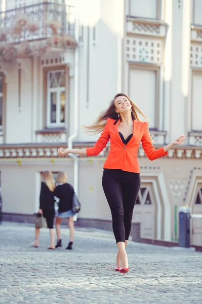 Modischer Look, Modell einer jungen Frau am heißen Tag, die in roter Jacke und schwarzer Hose durch die Stadt läuft, blonde Haare flattern über dem warmen Hintergrund der Stadt — Stockfoto