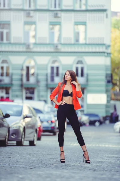 Fashionable look, hot day model of a young woman walking in the city, wearing a red jacket and black pants, blond hair outdoors over the city warm background — Stock Photo, Image