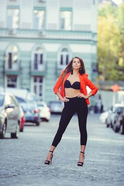 Modischer Look, Modell einer jungen Frau am heißen Tag, die in roter Jacke und schwarzer Hose durch die Stadt läuft, blonde Haare über dem warmen Hintergrund der Stadt — Stockfoto