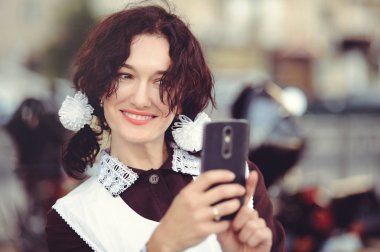 Güzel genç kadın, kıvırcık saç modeli ve açık havada yay. Mutlu ve sağlıklı bir Sovyet okul üniforması giymiş. Smartphone'da çıkartıyor. Nostalji kavramı