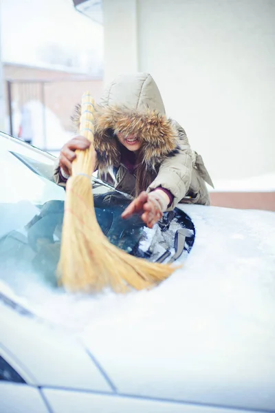 Счастливая девушка убирает свою машину со снега в городе. Рядом с метлой — стоковое фото