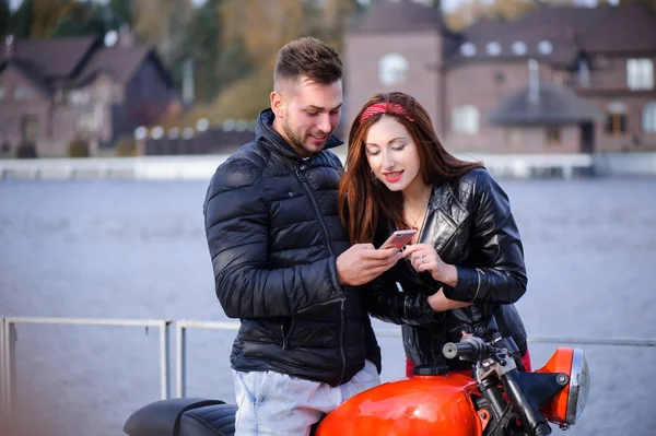 Casal elegante e na moda no amor olhar para o telefone em uma moto flerta close-up em um fundo do final do outono no parque — Fotografia de Stock