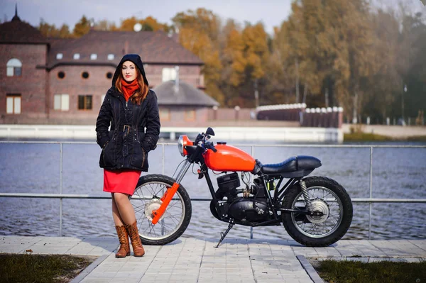 Joven mujer bonita en una capucha en la estación fría contra una motocicleta roja y bosque amarillo, pelo rojo y una bufanda alrededor de su cuello — Foto de Stock