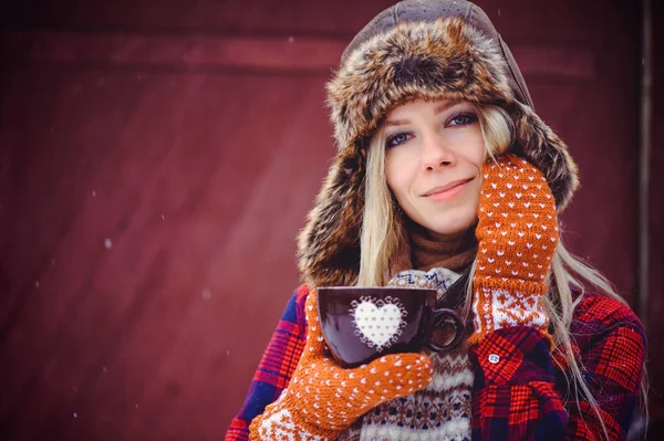 Mulher gentil em um chapéu de inverno e varejistas com uma xícara de chá quente ou café em um fundo vermelho, uma filmagem de publicidade brilhante — Fotografia de Stock