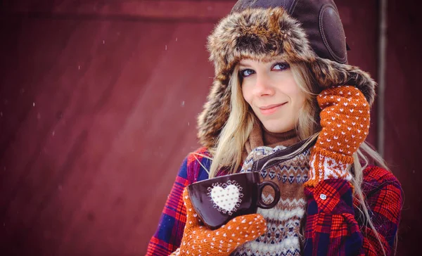 Mulher gentil em um chapéu de inverno e varejistas com uma xícara de chá quente ou café em um fundo vermelho, uma filmagem de publicidade brilhante — Fotografia de Stock