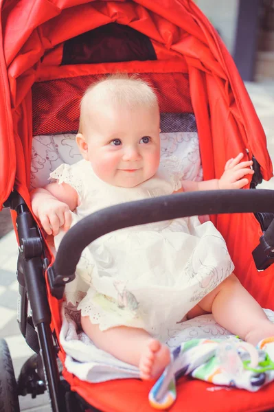 Niño de medio año en un cochecito, una niña con un vestido blanco en un cochecito rojo se regocija. Concepto educación de los niños, bienes de los niños —  Fotos de Stock