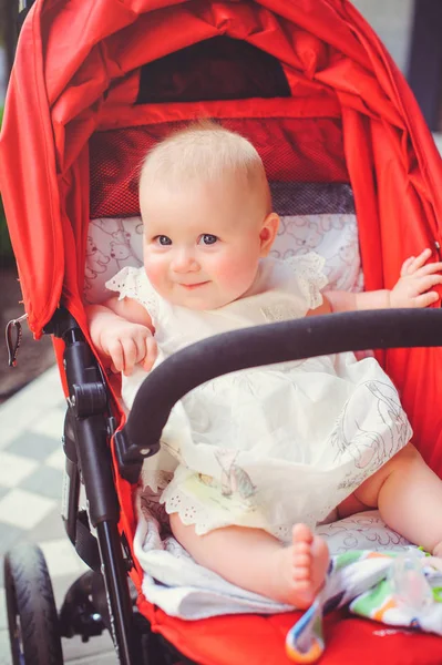 Niño de medio año en un cochecito, una niña con un vestido blanco en un cochecito rojo se regocija. Concepto educación de los niños, bienes de los niños —  Fotos de Stock