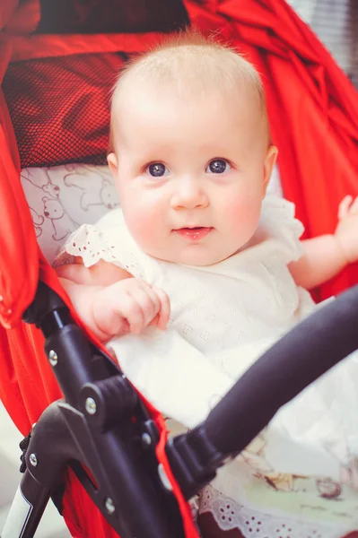 Niño de medio año en un cochecito, una niña con un vestido blanco en un cochecito rojo se regocija. Concepto educación de los niños, bienes de los niños —  Fotos de Stock