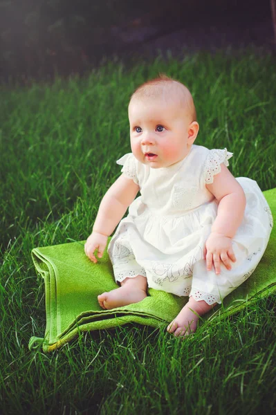 一个半岁的孩子坐在院子里的草地上, 穿着一件白色的连衣裙, 欣喜地, 6 月。儿童、儿童用品的概念教育 — 图库照片