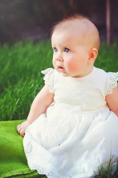 Niño de medio año sentado en la hierba en el patio, vestido con un vestido blanco se regocija, 6 meses. Concepto educación de los niños, bienes de los niños —  Fotos de Stock