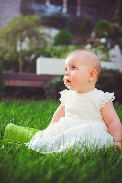 Полугодовалый ребенок, сидящий на траве во дворе, одетый в белое платье, радуется, 6 месяцев. Концепция воспитания детей, товаров для детей — стоковое фото