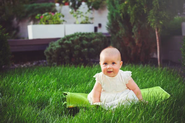 Half-jaar oude kind zittend op het gras in de tuin, gekleed in een witte jurk verheugt zich, 6 maanden. Concept onderwijs voor kinderen, kinderen van goederen — Stockfoto