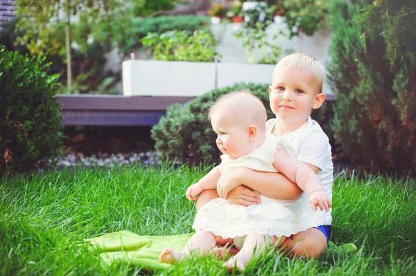 Gelukkig spelen broertje knuffels zijn zus Babysitting op gras in een groene tuin, een concept van liefde en ouderschap — Stockfoto