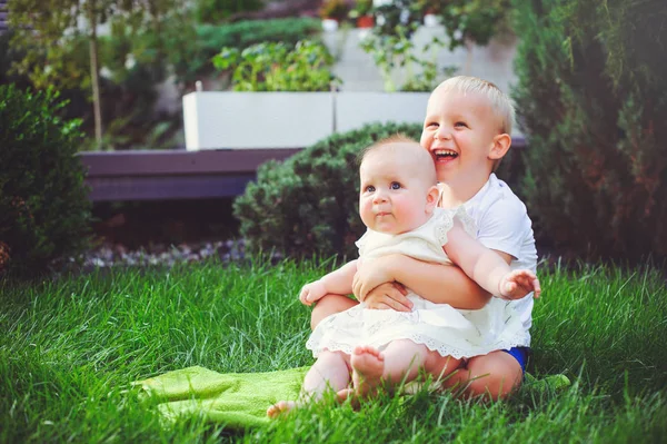 Glada lilla bror spela kramar sin syster baby sitter på gräset i en grön trädgård, begreppet kärlek och föräldraskap — Stockfoto
