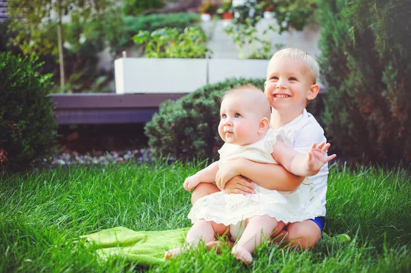Adik kecil yang bahagia bermain pelukan adiknya bayi duduk di rumput di kebun hijau, konsep cinta dan pengasuhan — Stok Foto