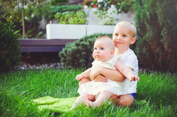 Boldog kis testvér játék hugs a húga ül a fűben, egy zöld kert, a szeretet és a nevelési koncepció baba — Stock Fotó