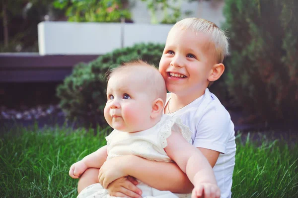 happy little brother playing hugs his sister baby sitting on grass in a green garden, concept of love and parenting