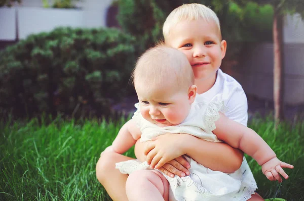 Adik kecil yang bahagia bermain pelukan adiknya bayi duduk di rumput di kebun hijau, konsep cinta dan pengasuhan — Stok Foto
