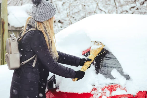 Winter portrait of a young woman cleaning snow from a car. Beauty blonde Model Girl laughs and cheerfully cleans the snow. Beautiful young woman outdoors