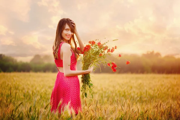 Bela mulher em um vestido vermelho com um buquê de papoilas em um campo de trigo ao pôr do sol, tonificação quente, felicidade e um estilo de vida saudável — Fotografia de Stock