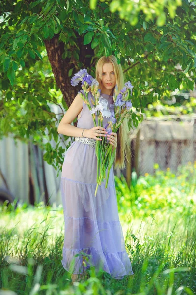 Attraktives junges Mädchen mit blonden Haaren und natürlichem Make-up, das blau lila Irisblüten auf einem Hintergrund im Freien riecht, Zartheit und Weichheit auf einem Naturhintergrund — Stockfoto