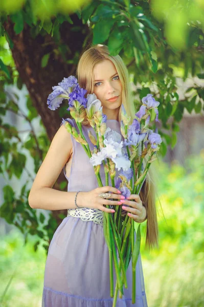 Muchacha joven atractiva con cabello rubio y maquillaje natural oliendo flores de iris azul púrpura sobre un fondo al aire libre, ternura y suavidad sobre un fondo natural — Foto de Stock