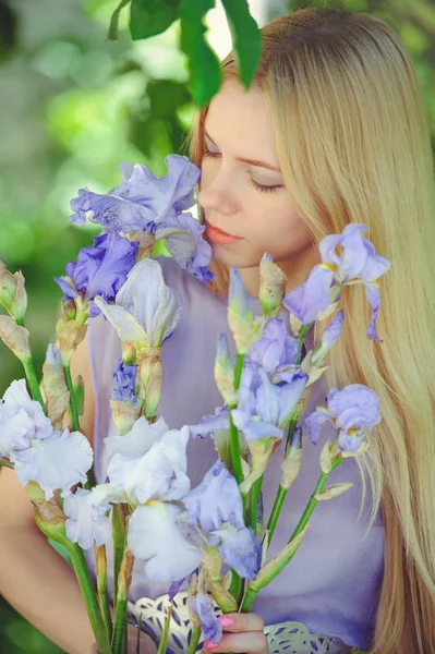 Rapariga atraente com cabelo loiro e maquiagem natural cheirando flores de íris roxas azuis em um fundo ao ar livre, ternura e maciez em um fundo da natureza — Fotografia de Stock