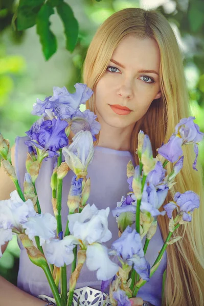 Aantrekkelijke jonge meisje met blonde dren haar en natuurlijke make-up ruiken blauw paarse iris bloemen op een achtergrond in openlucht, tederheid en zachtheid op een natuur-achtergrond — Stockfoto