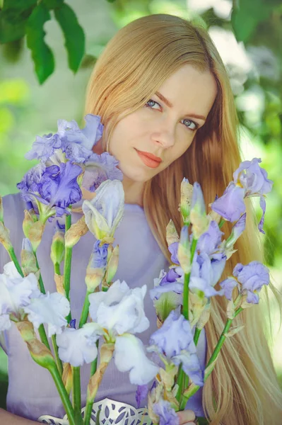 Muchacha joven atractiva con cabello rubio y maquillaje natural oliendo flores de iris azul púrpura sobre un fondo al aire libre, ternura y suavidad sobre un fondo natural — Foto de Stock