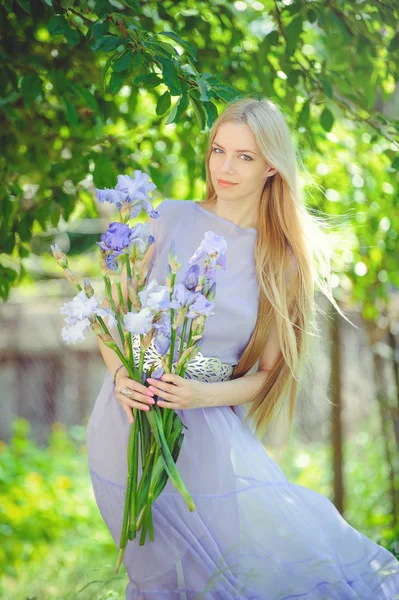 有吸引力的年轻女孩与金发儿歌头发和自然化妆嗅到的蓝色紫色虹膜花在背景户外, 柔情和柔软的自然背景 — 图库照片