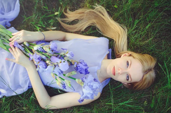 Muchacha joven atractiva con cabello rubio y maquillaje natural oliendo flores de iris azul púrpura tumbadas en la hierba al aire libre, ternura y suavidad en el fondo de la naturaleza — Foto de Stock