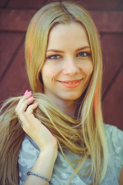 Attractive modest young blond woman playing with hair on red wooden background her hair painted in pink striped striped, in blue dress shy and inquisitive