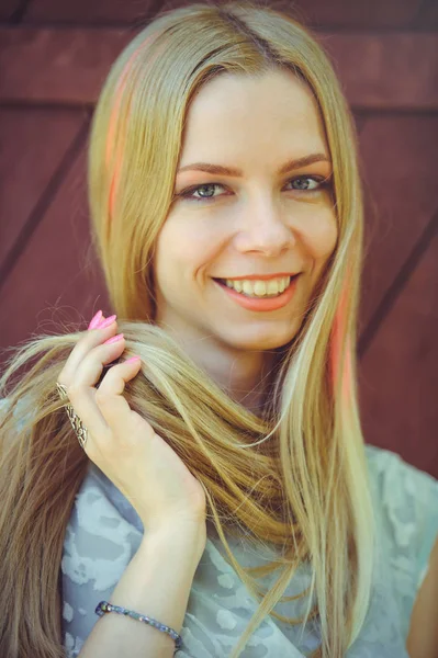 Atrakcyjne skromna młoda blond kobieta grając z włosów na czerwonym tle drewniane malowane jej włosy w różowe paski paski, w niebieskiej sukience nieśmiały i ciekawy — Zdjęcie stockowe