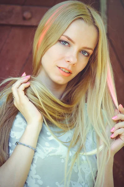 Ελκυστική μέτρια νεαρή γυναίκα ξανθιά παίζει με τα μαλλιά σε κόκκινο φόντο ξύλινη τα μαλλιά της βαμμένα σε ροζ ριγέ ριγέ, σε μπλε φόρεμα ντροπαλός και περίεργο — Φωτογραφία Αρχείου
