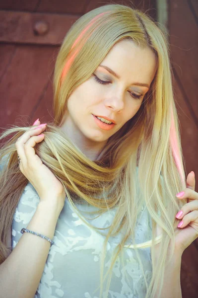 Attractive modest young blond woman playing with hair on red wooden background her hair painted in pink striped striped, in blue dress shy and inquisitive