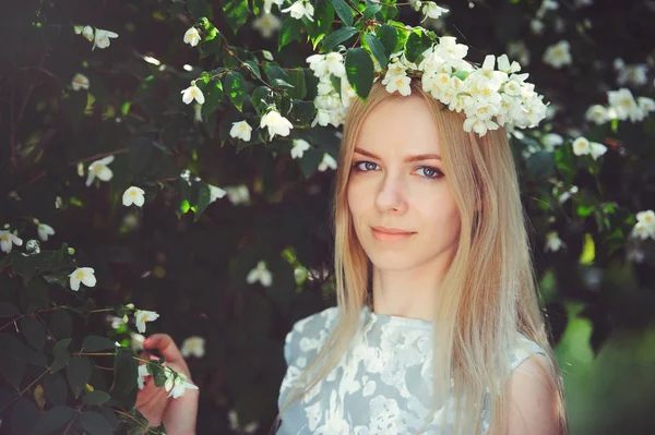 Attractive modest young girl with blonde with jasmine flowers wreath on head long hair and natural make-up in white dress outdoors, tenderness and softness on nature background