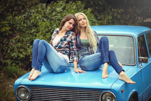 Cheerful Attractive two young blonde girl and brunette posing on the hood of an old rusty car, dressed in jeans and shirts on a nature background — Stock Photo, Image