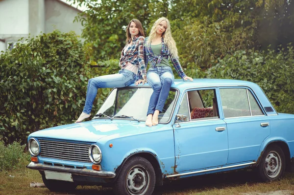 Cheerful Attractive two young blonde girl and brunette posing on the hood of an old rusty car, dressed in jeans and shirts on a nature background — Stock Photo, Image