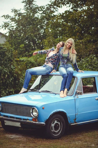 Cheerful Attractive two young blonde girl and brunette posing on the hood of an old rusty car, dressed in jeans and shirts on a nature background — Stock Photo, Image