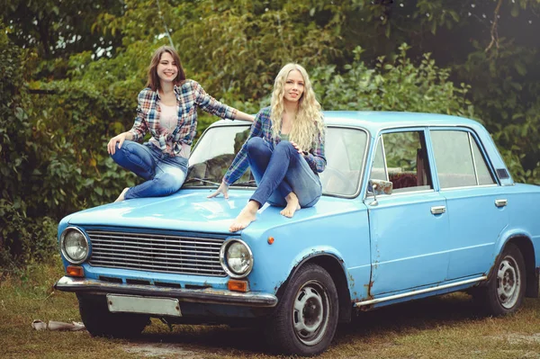 Cheerful Attractive two young blonde girl and brunette posing on the hood of an old rusty car, dressed in jeans and shirts on a nature background — Stock Photo, Image