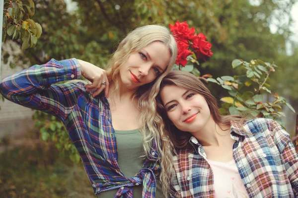 Alegre atractivo dos chica rubia joven y morena posando sobre fondo de la naturaleza, con pantalones vaqueros y camisas, hermanas —  Fotos de Stock